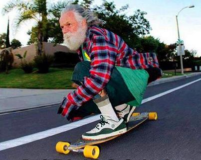 Un señor mayor con barba blanca montando en monopatin por un carretera